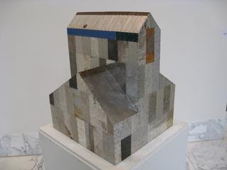 A small house sculpture on a white pedestal in an art gallery with marble flooring.