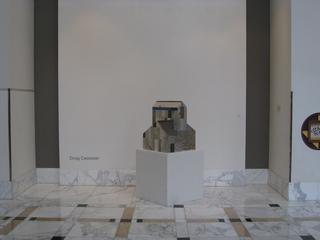 A small house sculpture by Doug Casebeer on a white pedestal in an art gallery with marble flooring.